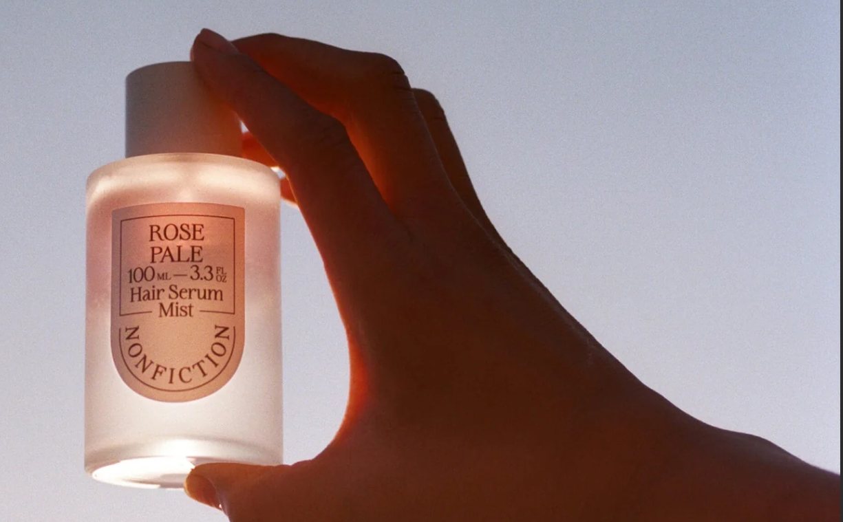 A hand holding the Rose Pale Hair Serum Mist bottle up to the light against a soft lilac sky.